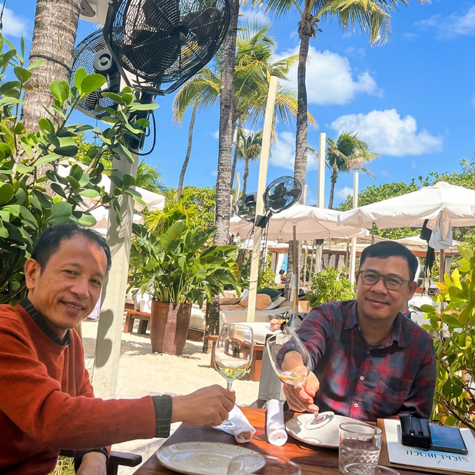 Two men toast with wine at an outdoor tropical restaurant surrounded by palm trees, lush plants, umbrellas, and cooling fans.