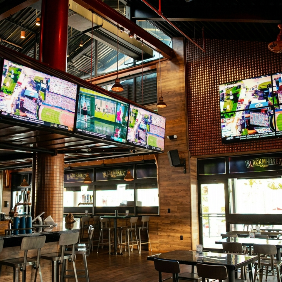 Industrial sports bar interior with numerous televisions showing live sports, pendant lighting, high-top tables, and Blackmarket branding.
