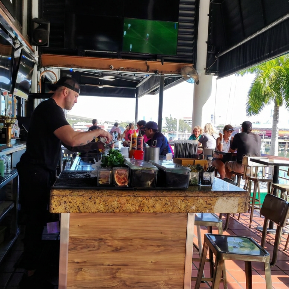 Bartender prepares drinks at a waterfront bar with high-top seating, sports televisions, palm trees, and marina views.