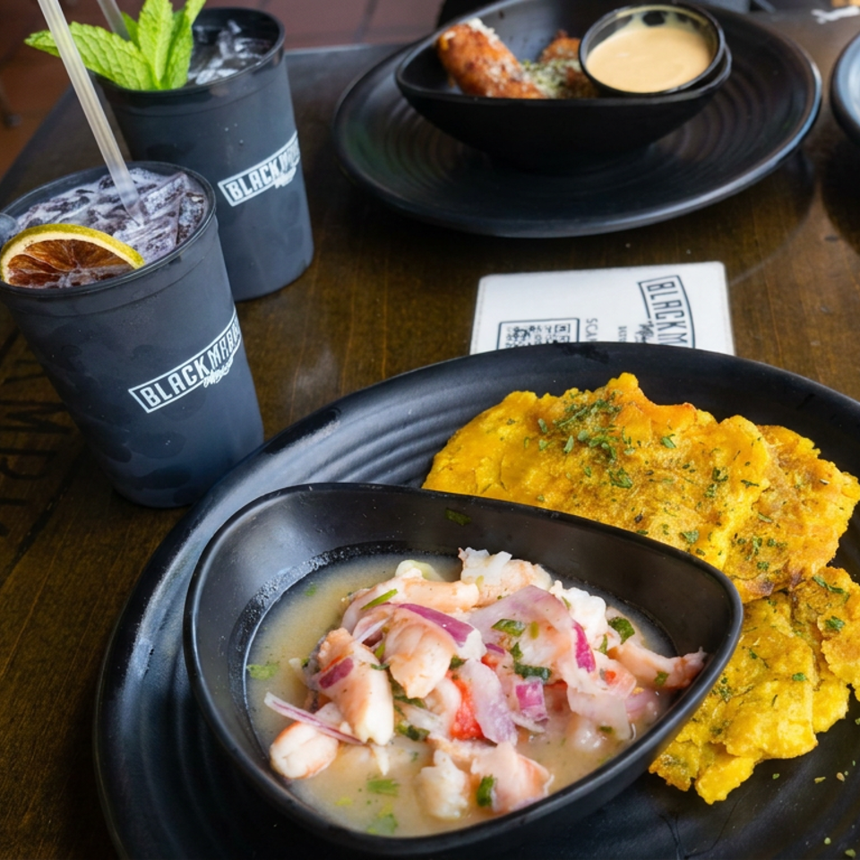 Black plates hold ceviche with red onion and herbs alongside golden fried plantains, with citrus cocktails on a restaurant table.