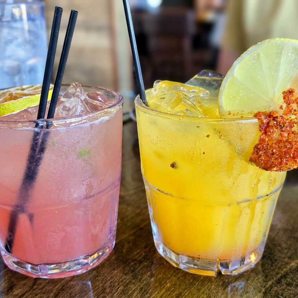 Two colorful iced cocktails sit on a wooden restaurant table, garnished with lime slices and a chili-salt rim.
