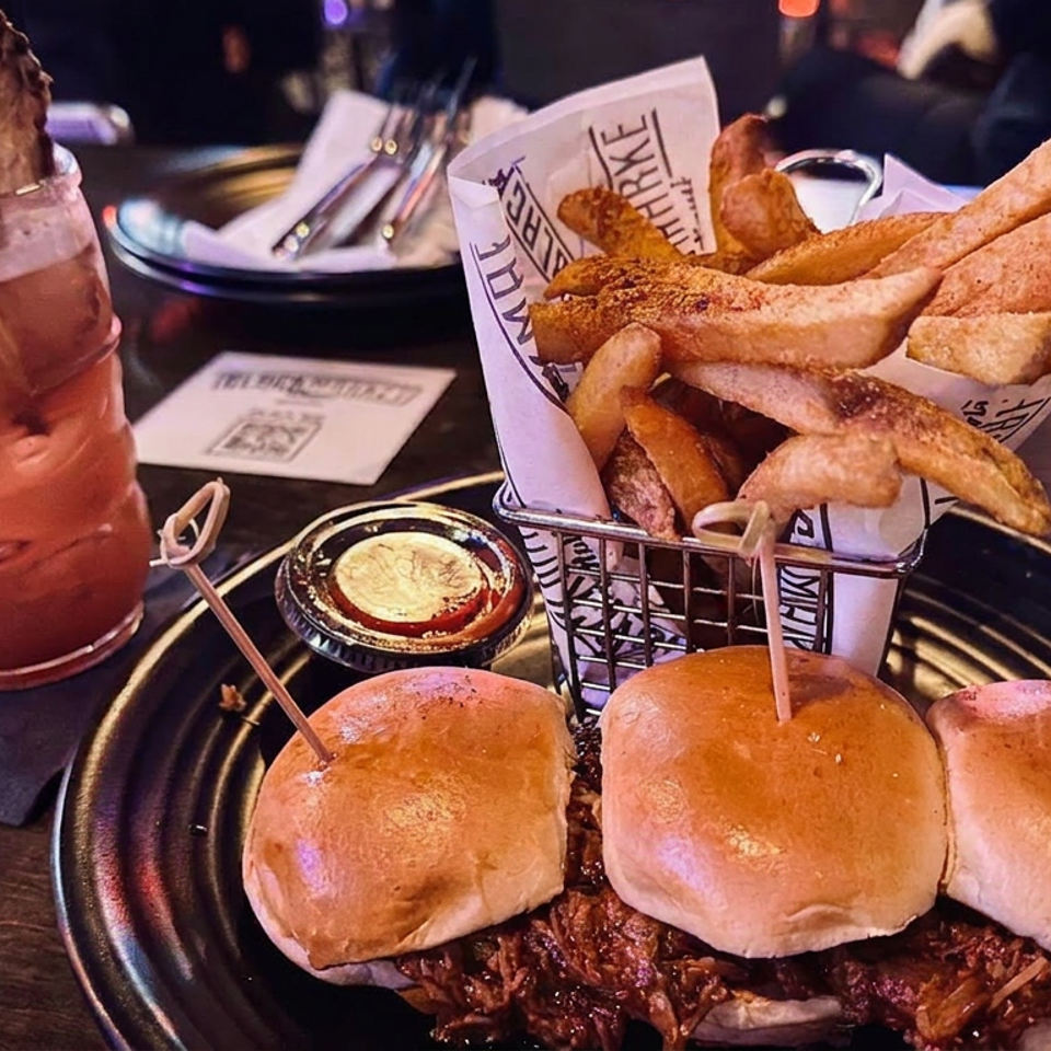 Restaurant platter with three pulled-beef sliders, fries, dipping sauce, and a mint-garnished red cocktail on a dark table.