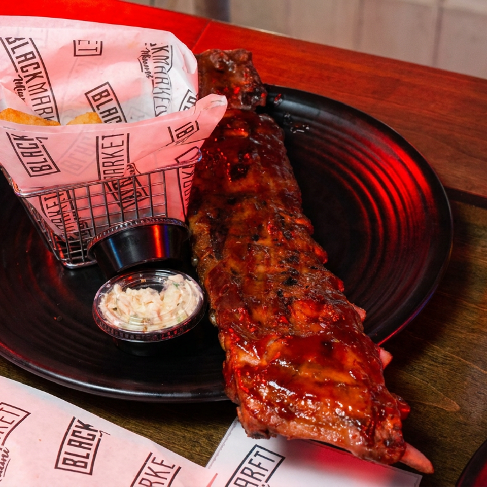 Glazed barbecue ribs served with fries, coleslaw, and a beer on a black plate at a wooden restaurant table.